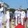 USS South Dakota (SSN 790) Holds Change of Command Ceremony A formal U.S. Navy ceremony is taking place on a pier or dock, featuring personnel in white dress uniforms. A senior officer wearing multiple service ribbons and medals is walking a red carpet while being saluted by a sideboy formation. The setting appears to be a naval installation with water and tree-covered hills visible in the background.