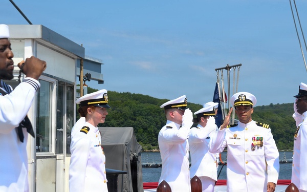 A formal U.S. Navy ceremony is taking place on a pier or dock, featuring personnel in white dress uniforms. A senior officer wearing multiple service ribbons and medals is walking a red carpet while being saluted by a sideboy formation. The setting appears to be a naval installation with water and tree-covered hills visible in the background.