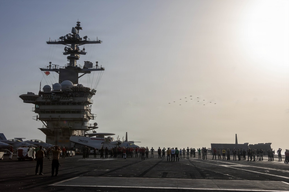 A large US Navy aircraft carrier flight deck is shown with crew members gathered and watching a formation flyover of multiple aircraft in the distance. The carrier's island superstructure is prominently visible on the left side, featuring radar domes, communication equipment, and multi-level command structures. The scene appears to depict a ceremonial or commemorative event given the assembled personnel and aerial formation.