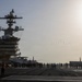 A large US Navy aircraft carrier flight deck is shown with crew members gathered and watching a formation flyover of multiple aircraft in the distance. The carrier's island superstructure is prominently visible on the left side, featuring radar domes, communication equipment, and multi-level command structures. The scene appears to depict a ceremonial or commemorative event given the assembled personnel and aerial formation.