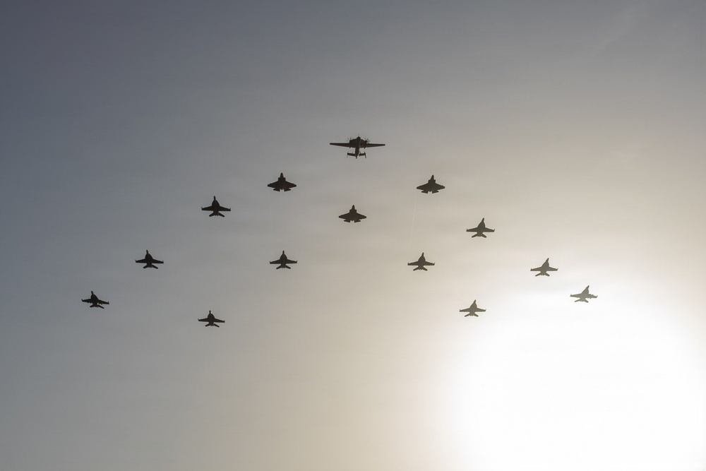 A large formation of military aircraft flying in a coordinated diamond or arrowhead pattern against a hazy, backlit sky. The formation includes what appears to be a mix of aircraft types, with a larger aircraft at the apex and numerous fighter jets arranged symmetrically behind it. The silhouette-style lighting makes precise identification difficult but suggests a formal flyover or commemorative aerial display.