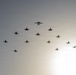 A large formation of military aircraft flying in a coordinated diamond or arrowhead pattern against a hazy, backlit sky. The formation includes what appears to be a mix of aircraft types, with a larger aircraft at the apex and numerous fighter jets arranged symmetrically behind it. The silhouette-style lighting makes precise identification difficult but suggests a formal flyover or commemorative aerial display.