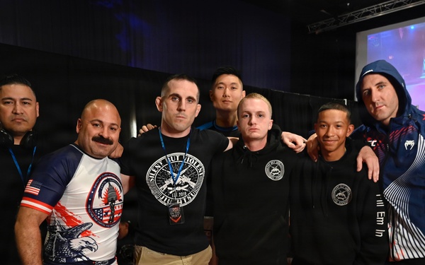 A group of approximately seven to eight individuals posing together on red competition mats at what appears to be a martial arts or grappling tournament. The participants are wearing a mix of rashguards, fight shorts, and athletic wear, suggesting they are competitors and coaches. A projection screen and indoor venue lighting are visible in the background.