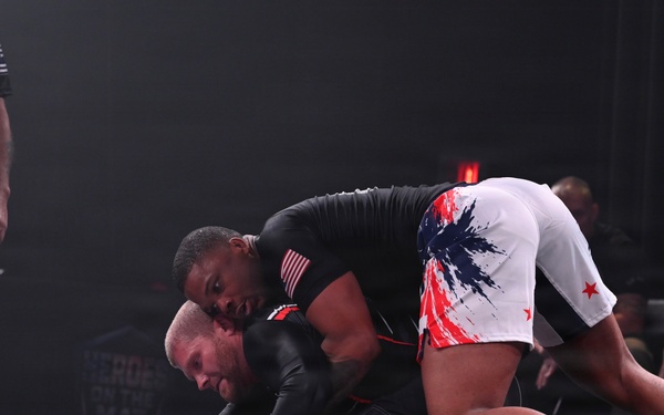 Two athletes are competing in a no-gi grappling/submission wrestling match at an event called 'Heroes on the Mat.' One competitor wearing white shorts with red and blue patriotic design has achieved a dominant top position over the opponent in all-black rash guard and shorts. A referee in a black 'Heroes on the Mat' shirt observes the match from the side.
