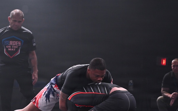 Two competitors are engaged in a grappling/submission wrestling match on a raised mat platform. A referee wearing a 'Heroes on the Mat' shirt closely monitors the action as one athlete in American flag-style shorts applies top pressure on a downed opponent. Spectators and corner staff are visible seated in the background to the right.