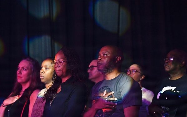 A group of approximately 8-10 adults standing at what appears to be a railing in a dimly lit venue, with several individuals placing their right hands over their hearts. The scene is illuminated by colorful stage lighting with purple, red, and blue tones. The gesture suggests they are participating in a national anthem or pledge of allegiance at a public event.