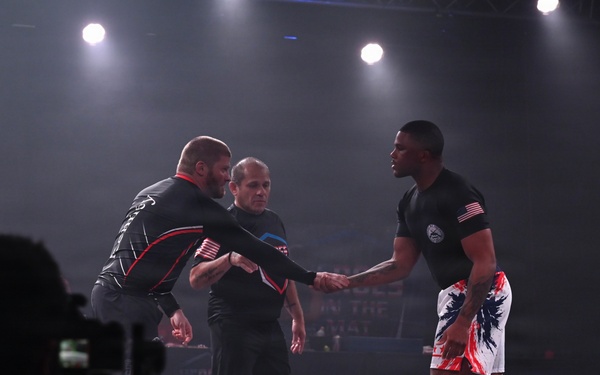 Two competitors are shaking hands on a grappling/submission wrestling mat under stage lighting, with a referee standing between them. Both athletes are wearing black rash guards and shorts, suggesting a no-gi grappling competition. A cameraman in silhouette is visible in the foreground capturing the pre-match or post-match handshake.