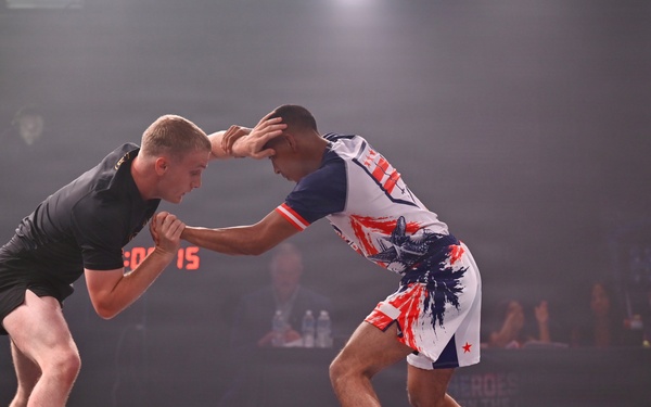 Two athletes compete in a no-gi grappling or submission wrestling match on a competition mat under bright stage lighting. One competitor wears all black shorts with a blue ankle band, while the other wears patriotic-themed white, red, and blue shorts with a red ankle band. The event appears to be a professional or semi-professional combat sports competition with spectators, officials, and a photographer visible in the background.