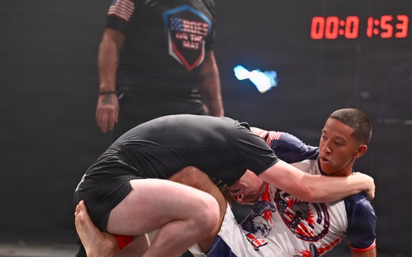 Two athletes are competing in a grappling/submission wrestling match on a gray competition mat. One competitor wearing a white and blue rash guard with red accents is seated on the mat while engaging with an opponent in a black rash guard and shorts. A referee in black stands in the background overseeing the match, with a digital scoreboard displaying '1:53' visible overhead.