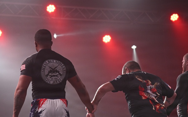 Three individuals stand on a stage facing away from the camera under dramatic red and white stage lighting. The setting appears to be a combat sports or martial arts event, suggested by the athletic attire and partial banner visible in the background reading 'HEROES'. One person appears to be supporting or guiding another, suggesting a post-match or ceremonial moment.