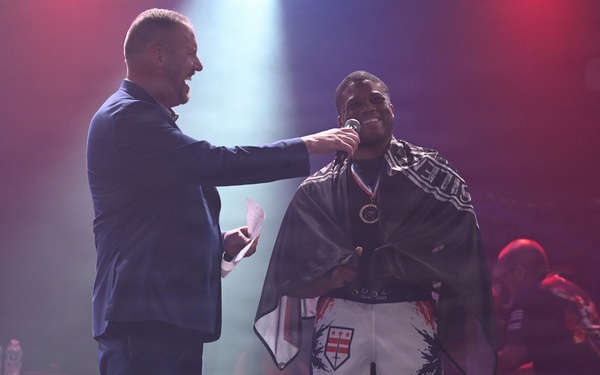 Two men are standing on a stage under colorful lighting, appearing to be at a combat sports or MMA event. One man in a blue suit is holding a microphone toward a fighter wearing white shorts with a shield/stars design and a dark cape or flag draped over his shoulders, along with a medal around his neck. The setting suggests a post-fight ceremony or award presentation.