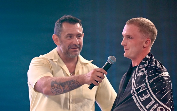 Two men are standing on a stage under blue-tinted lighting. One man in a light yellow short-sleeve shirt with visible arm tattoos is holding a microphone toward the other man, who is wearing a dark patterned cape or flag draped over his shoulders. The setting appears to be a formal event, ceremony, or competition presentation.