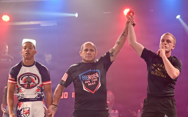 A martial arts or grappling competition victory ceremony is taking place on a lit stage. A referee wearing a 'Heroes on the Mat' shirt raises the right hand of the winner, who is dressed in black shorts and a black shirt from Cincinnati Martial Arts Academy. The defeated competitor stands to the left wearing a 'Guardians' team rash guard and white fight shorts.