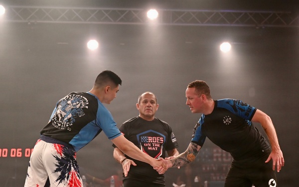 Two competitors are shaking hands at the start or end of a grappling/submission wrestling match, overseen by a referee standing between them. The event appears to be 'Heroes on the Mat,' a combat sports competition held in an indoor arena with professional lighting. A digital countdown clock is visible in the background showing '00:06:0x'.