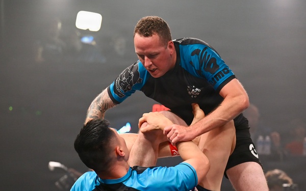 Two athletes are competing in a no-gi grappling or submission wrestling match on a competition mat under bright event lighting. The standing competitor appears to be attempting a leg lock or ankle submission on the grounded opponent. Both athletes are wearing matching blue rash guards, suggesting they may be from the same team or the attire is event-issued.