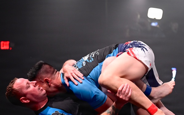 Two athletes are competing in a no-gi grappling or submission wrestling match on a dark mat under stage lighting. One competitor in a blue rash guard is on his back while the other in a black and blue rash guard with white shorts is on top, engaged in a ground fighting exchange. A digital timer on the right side of the image reads 00:05:20, indicating an active timed competition.