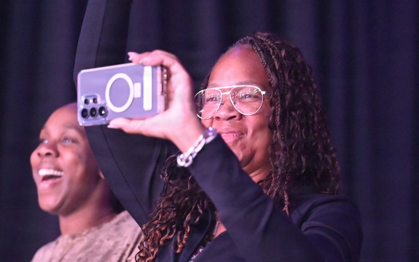 Three individuals are captured at what appears to be a live event or performance venue with a dark curtain backdrop. The central figure, wearing glasses and a black hoodie, is holding up a smartphone to record or photograph the event. The group appears to be enjoying themselves, with visible laughter and clapping.