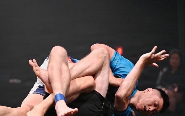 Two male athletes are engaged in a grappling/submission wrestling match on a competition mat. One competitor wearing a blue rashguard and black shorts appears to be applying a ground technique while the other, wearing white/red shorts, is entangled in the hold. Spectators are visible in the blurred background observing the match.