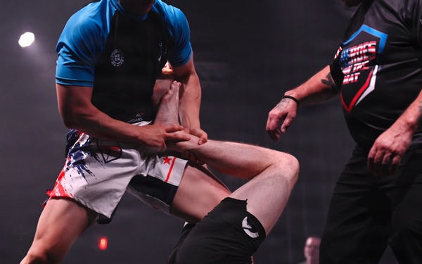 A grappling/submission wrestling match is taking place on a dark competition mat under stage lighting. One competitor in blue and white shorts is standing and applying a leg lock or ankle submission on a second competitor dressed in all black who is on the ground. A referee in a black official shirt with an American flag patch is observing the action closely.