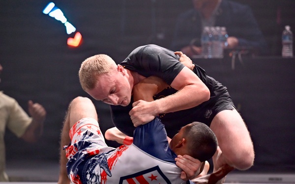 Two athletes are competing in a grappling/submission wrestling match on a gray mat. One competitor wearing a black rash guard is on top, applying pressure to an opponent wearing a patriotic red, white, and blue uniform. The event appears to be 'Heroes on the Mat,' a competition likely involving military or veteran athletes.