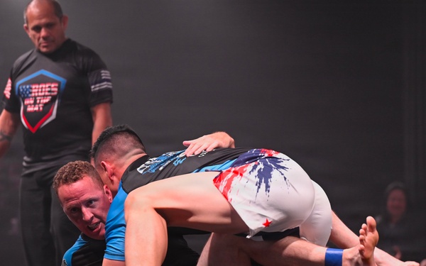 A grappling/submission wrestling match is taking place on a raised mat under stage lighting. Two competitors are engaged in ground fighting, with one athlete in a blue rash guard on the bottom and another in white-and-red shorts on top. A referee in a black 'Heroes on the Mat' shirt stands over them, observing the action closely.