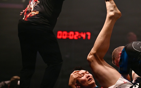 A grappling/submission wrestling match is taking place on a dark mat. One competitor in a blue rash guard is on his back with his leg raised, while an opponent in a black rash guard applies pressure from above. A referee in a black graphic t-shirt and black pants stands nearby overseeing the match, with a digital timer in the background reading 00:02:47.