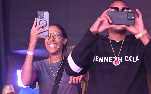 A group of people at what appears to be a live event or concert, enthusiastically recording with their smartphones. Two individuals stand prominently in the foreground — a smiling woman in glasses holding up an iPhone and a man in a Kenneth Cole hoodie filming with his phone. The venue has purple/blue ambient lighting suggesting an indoor entertainment setting.