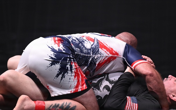 Two athletes are competing in a grappling/submission wrestling match on a dark mat surface. One competitor wearing a white and red/blue eagle-print rash guard is on top, while the other in a black rash guard with a US flag patch is on the bottom in a defensive position. The match appears to be a no-gi submission grappling competition.