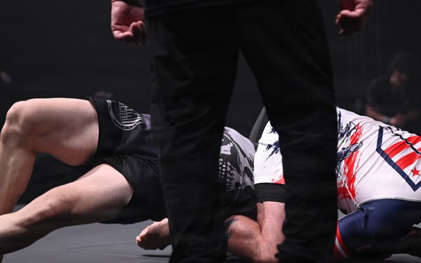 A grappling or MMA competition is taking place on a dark mat surface. Two athletes are engaged in ground fighting, with one competitor in a white patriotic-themed rashguard appearing to be in a defensive position on the mat. A referee in black pants and shoes stands over them, monitoring the action closely.