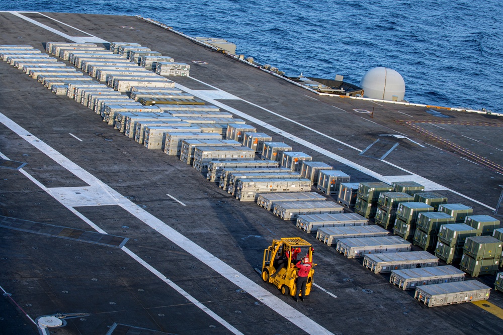 An aerial view of a naval vessel's flight deck, likely an aircraft carrier or amphibious assault ship, covered with numerous rows of military supply containers and cargo boxes. A yellow forklift operated by a crew member in a red jersey is actively moving among the organized rows of containers. The ocean is visible in the background, and a white radome (satellite communication dome) is visible on the ship's deck edge.