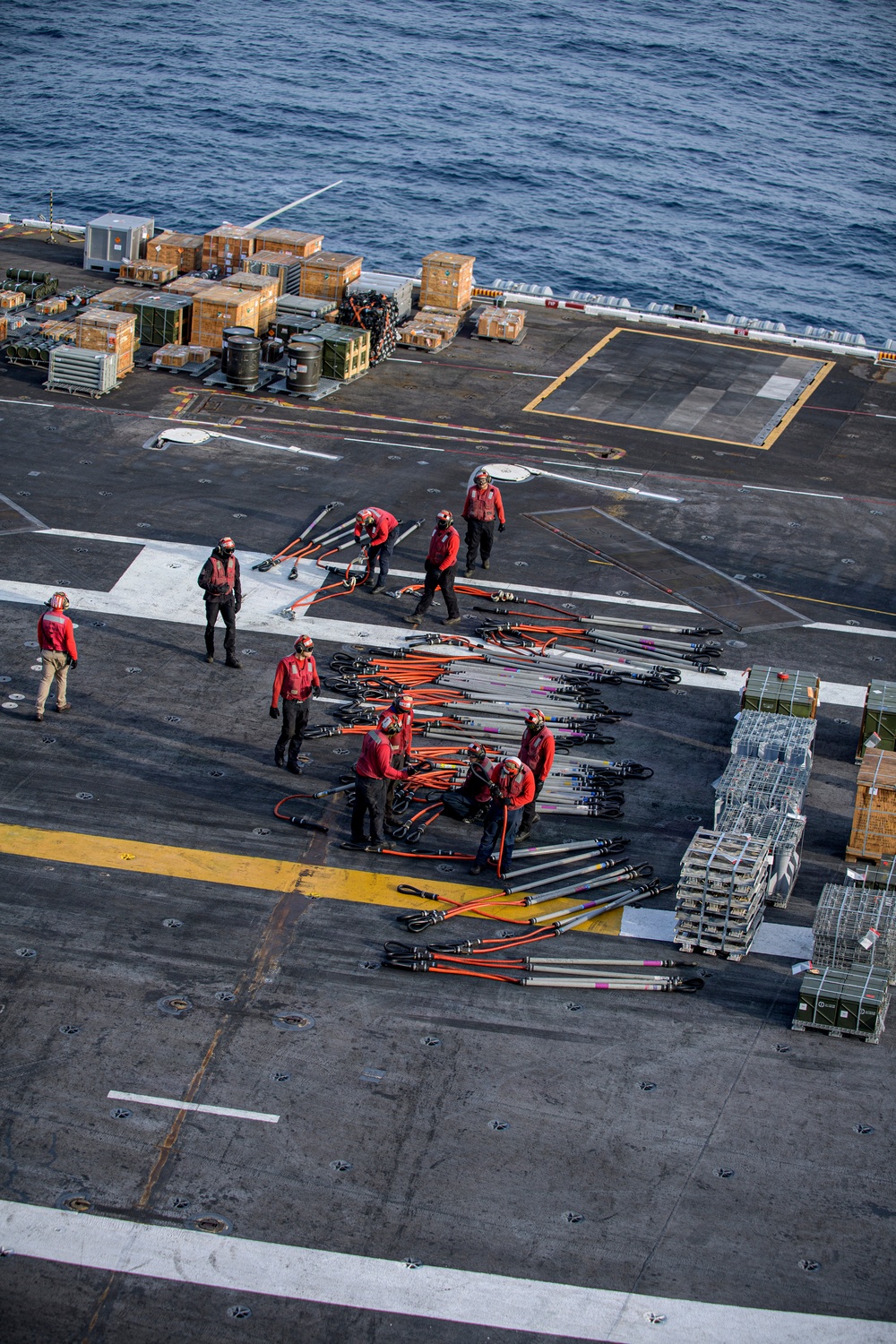 An aerial photograph taken from above a naval aircraft carrier flight deck, showing crew members in red jerseys handling what appear to be cargo straps or tie-down chains. The deck is populated with numerous wooden crates, pallets, metal containers, and wire cages, suggesting an underway replenishment or cargo transfer operation. The ocean is visible in the upper portion of the image, confirming the vessel is at sea.
