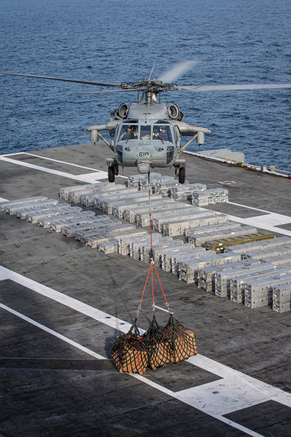 A U.S. Navy helicopter numbered '610', consistent with an MH-60S Seahawk, hovers over a ship's flight deck performing a vertical replenishment (VERTREP) operation. The helicopter is lifting a cargo net loaded with supplies using an external sling load with orange straps. The flight deck is densely packed with numerous silver/gray military supply containers arranged in organized rows.