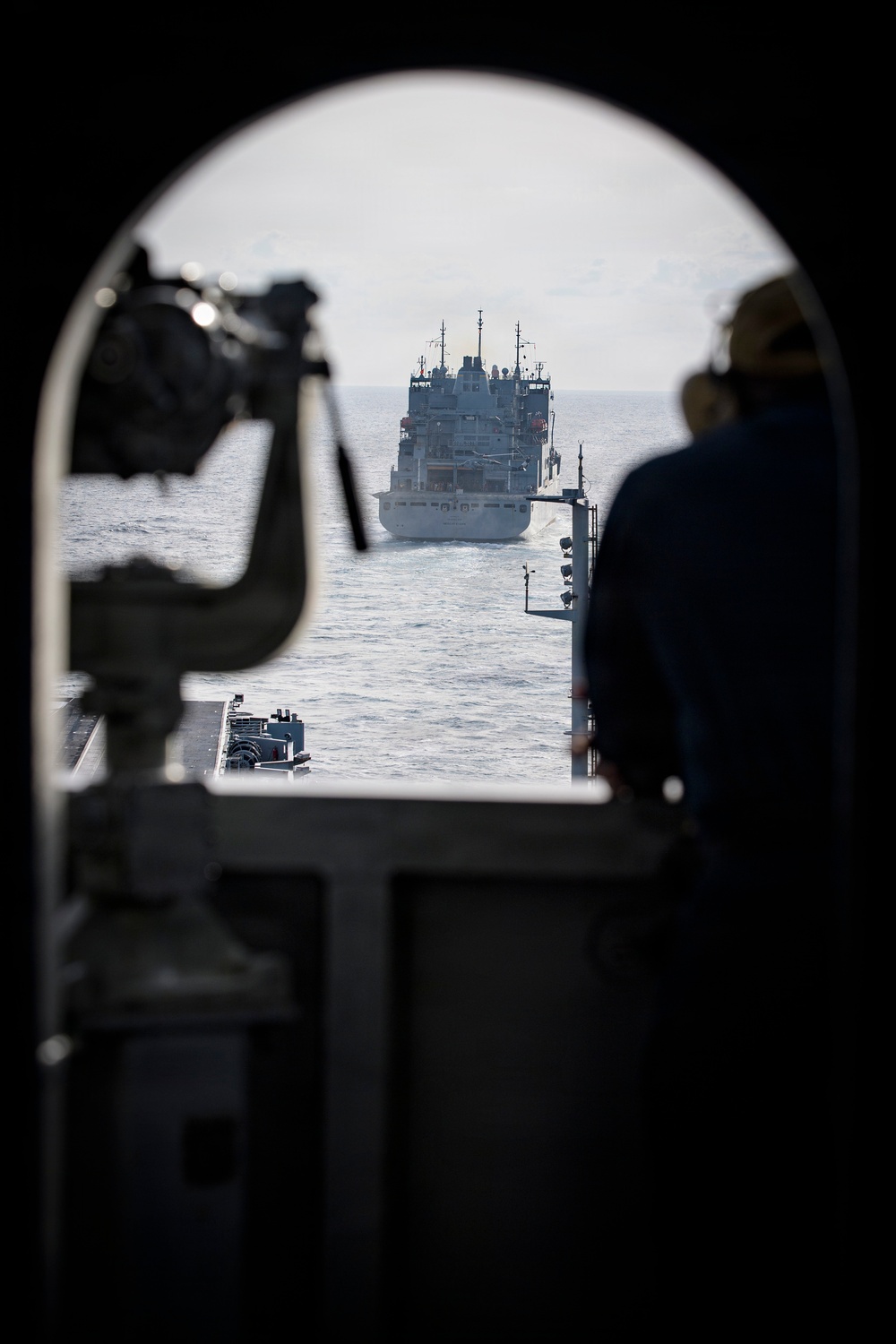 The image is taken from inside a naval vessel looking through an arched porthole or hatch opening toward an open ocean. A large auxiliary or replenishment ship is visible ahead in the water, suggesting a potential underway replenishment (UNREP) or formation sailing scenario. A silhouetted crew member stands on the right side observing the vessel ahead.