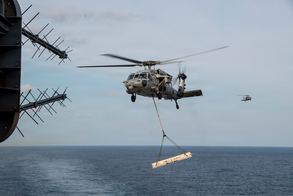 A military helicopter, identified as number 610 and consistent with an MH-60 Seahawk, is performing a vertical replenishment (VERTREP) operation over open ocean. The helicopter is carrying a cargo pallet suspended by a sling load beneath it, approaching what appears to be a naval vessel. A second helicopter is visible in the background, suggesting a coordinated logistics operation.