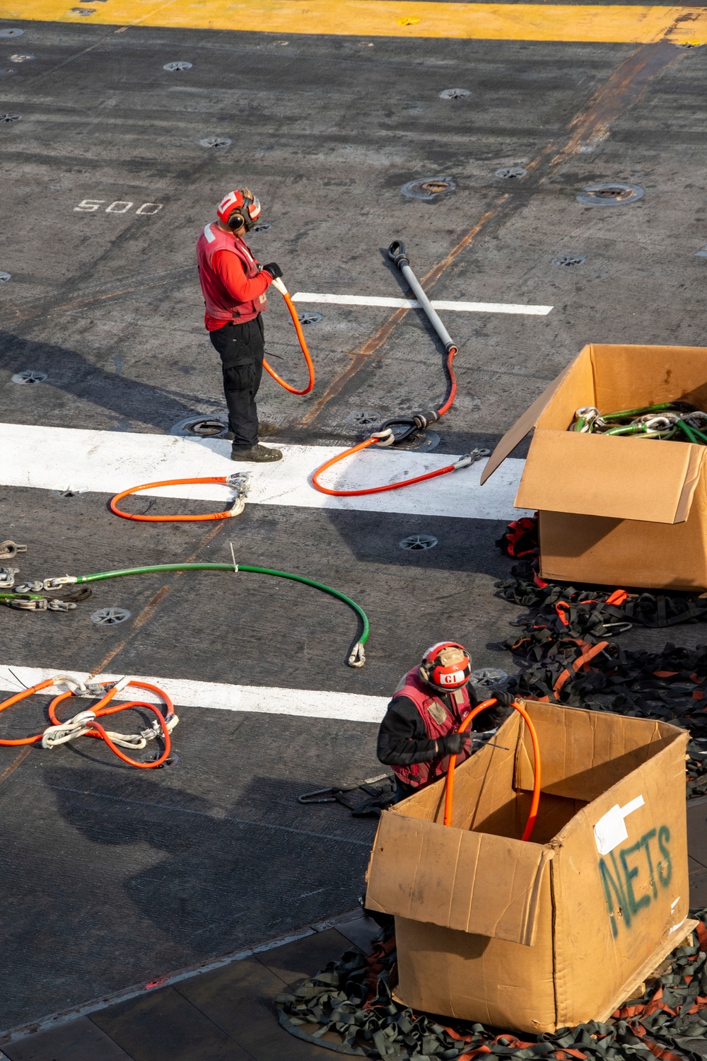 Two personnel in red jerseys and cranial helmets are working on what appears to be a naval aircraft carrier flight deck, handling orange and green safety lanyards or tie-down straps. They are organizing equipment near large cardboard boxes, one of which is labeled 'NETS'. The number '500' is painted on the deck surface, consistent with carrier deck markings.