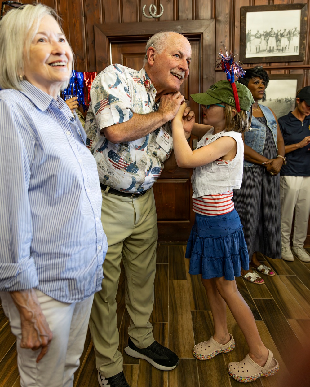 A festive indoor gathering with patriotic American themes, likely a Fourth of July celebration. An elderly man wearing an American flag-print shirt is interacting warmly with a young girl dressed in red, white, and blue, while an older woman stands nearby smiling. Several other guests are visible in the background of what appears to be a wood-paneled room.