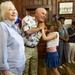 A festive indoor gathering with patriotic American themes, likely a Fourth of July celebration. An elderly man wearing an American flag-print shirt is interacting warmly with a young girl dressed in red, white, and blue, while an older woman stands nearby smiling. Several other guests are visible in the background of what appears to be a wood-paneled room.