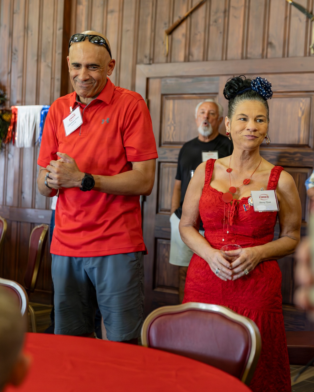 Two individuals wearing red outfits and name badges stand together in what appears to be a formal gathering or event in a wood-paneled room. The woman on the right wears a red dress with a matching necklace and has her hair styled up with a blue polka-dot accessory, while the man on the left wears a red polo shirt with sunglasses on his head. A third person is visible in the background wearing a black shirt with a name badge.
