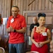 Two individuals wearing red outfits and name badges stand together in what appears to be a formal gathering or event in a wood-paneled room. The woman on the right wears a red dress with a matching necklace and has her hair styled up with a blue polka-dot accessory, while the man on the left wears a red polo shirt with sunglasses on his head. A third person is visible in the background wearing a black shirt with a name badge.