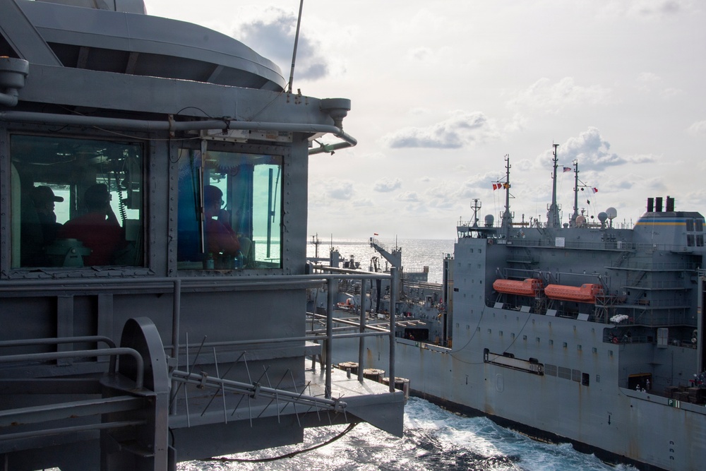 Two naval vessels are conducting an underway replenishment (UNREP) operation at sea, with hoses and lines connecting the ships while both are underway. The image is taken from the bridge wing of one vessel looking toward a larger auxiliary replenishment ship alongside. Personnel are visible inside the bridge structure observing the operation.