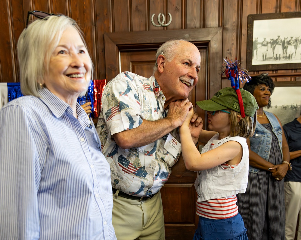 A cheerful patriotic gathering, likely a Fourth of July or American holiday celebration, taking place in a wood-paneled room. An elderly man wearing an American flag-themed shirt is interacting playfully with a young girl dressed in red, white, and blue with a festive hat. Several other adults are present in the background, all appearing to enjoy the occasion.