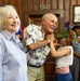 A cheerful patriotic gathering, likely a Fourth of July or American holiday celebration, taking place in a wood-paneled room. An elderly man wearing an American flag-themed shirt is interacting playfully with a young girl dressed in red, white, and blue with a festive hat. Several other adults are present in the background, all appearing to enjoy the occasion.