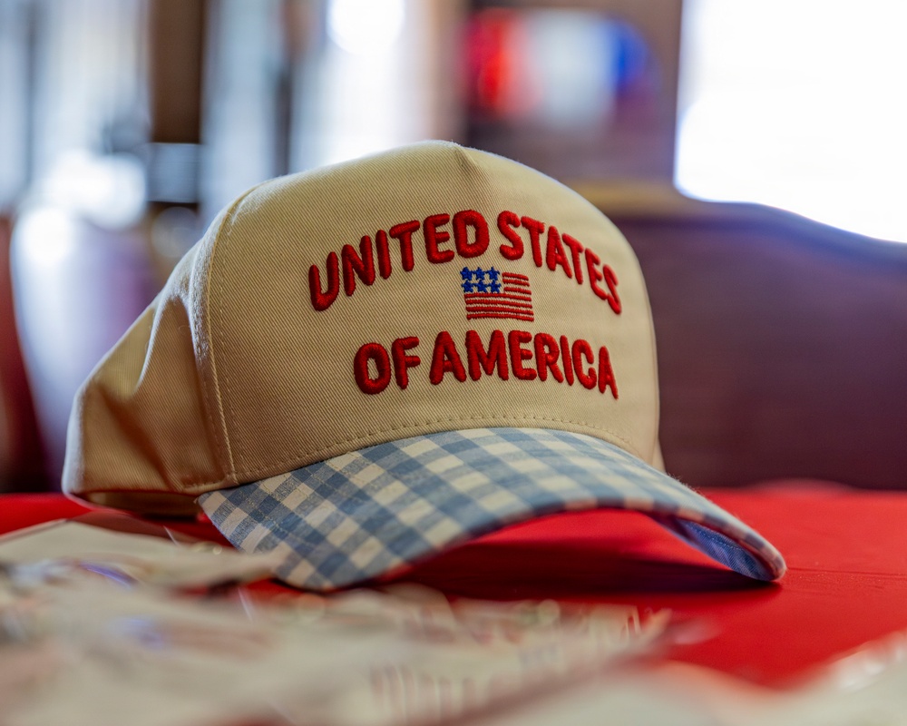 A khaki/tan baseball cap embroidered with 'United States of America' and a small American flag graphic is displayed on a red surface. The cap features a distinctive blue and white gingham/plaid patterned brim. The image is a close-up product-style photograph taken in what appears to be an indoor setting.