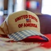 A khaki/tan baseball cap embroidered with 'United States of America' and a small American flag graphic is displayed on a red surface. The cap features a distinctive blue and white gingham/plaid patterned brim. The image is a close-up product-style photograph taken in what appears to be an indoor setting.