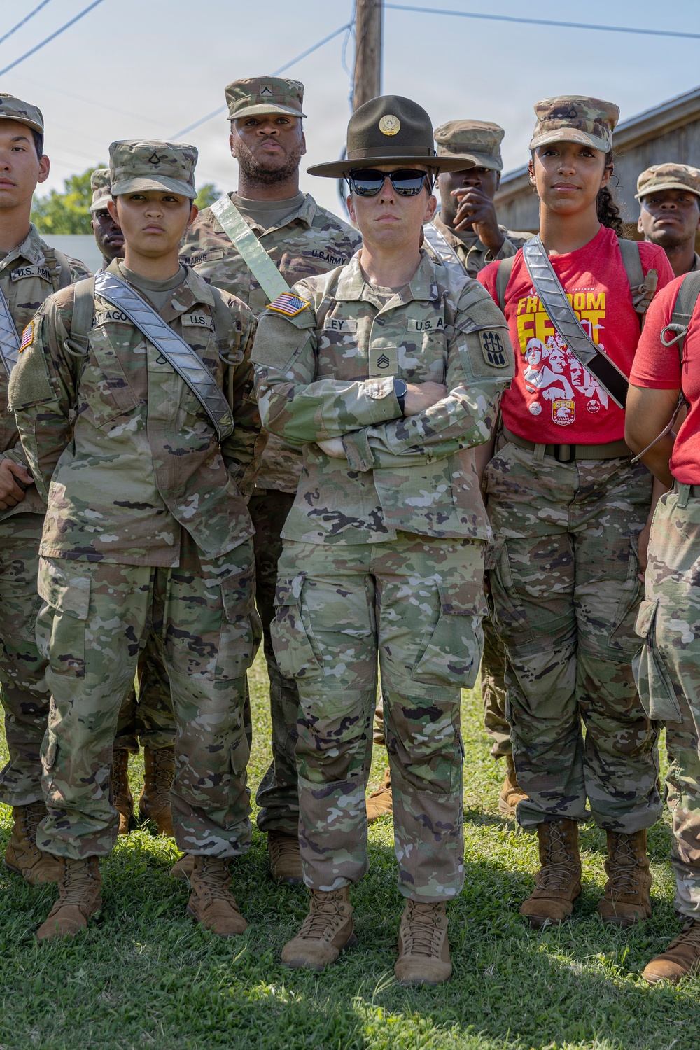 A group of U.S. Army soldiers standing at attention outdoors on grass. The central figure is a Drill Sergeant identifiable by the distinctive campaign/drill sergeant hat (Smokey Bear hat) and sunglasses, with arms crossed. Surrounding personnel wear OCP (Operational Camouflage Pattern) uniforms with various rank insignia and sashes.