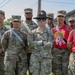 A group of U.S. Army soldiers standing at attention outdoors on grass. The central figure is a Drill Sergeant identifiable by the distinctive campaign/drill sergeant hat (Smokey Bear hat) and sunglasses, with arms crossed. Surrounding personnel wear OCP (Operational Camouflage Pattern) uniforms with various rank insignia and sashes.