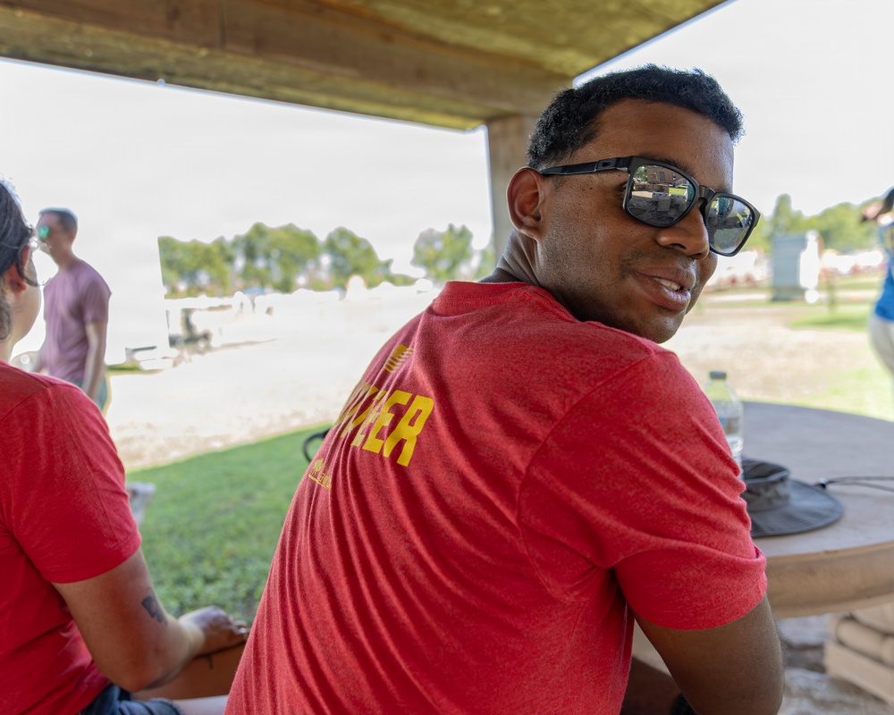 A man wearing a red t-shirt with yellow lettering and dark sunglasses is seated under a covered outdoor structure, turning to look back toward the camera. The setting appears to be a recreational outdoor area near a body of water or beach. Several other people are visible in the background in a park-like environment.