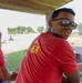 A man wearing a red t-shirt with yellow lettering and dark sunglasses is seated under a covered outdoor structure, turning to look back toward the camera. The setting appears to be a recreational outdoor area near a body of water or beach. Several other people are visible in the background in a park-like environment.