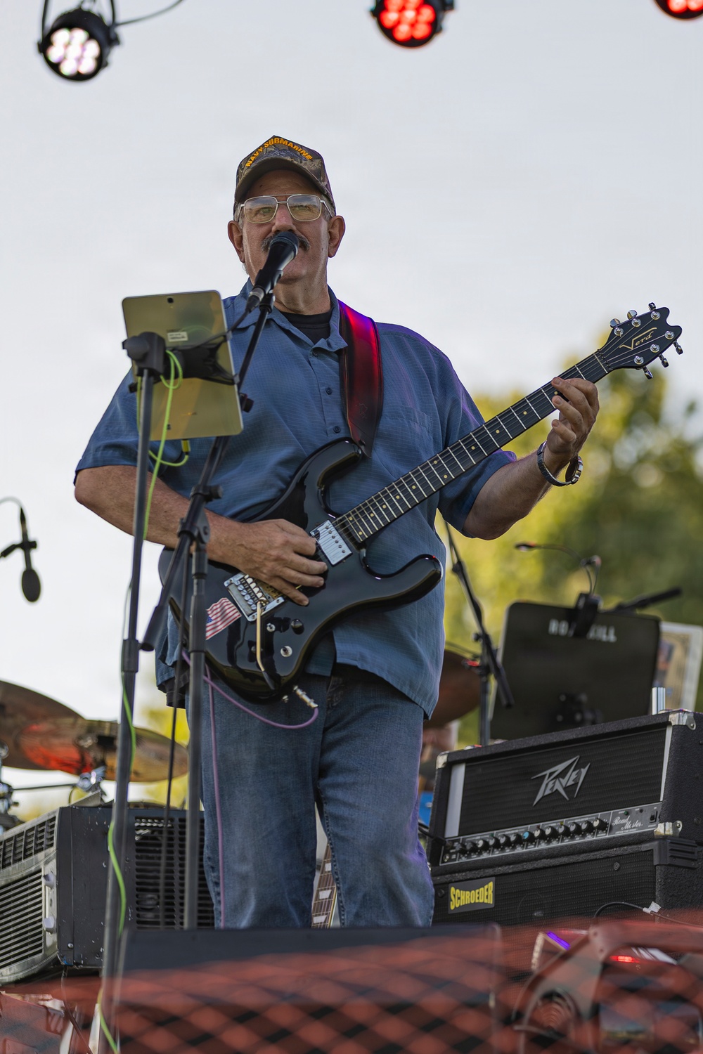 A middle-aged male guitarist is performing on an outdoor stage, singing into a microphone while playing a black electric guitar. He is wearing a blue button-up shirt, jeans, glasses, and a cap with text reading 'Navy Submarine'. Stage equipment including a Peavey amplifier, a Schroeder cabinet, and stage lighting is visible in the background.