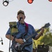 A middle-aged male guitarist is performing on an outdoor stage, singing into a microphone while playing a black electric guitar. He is wearing a blue button-up shirt, jeans, glasses, and a cap with text reading 'Navy Submarine'. Stage equipment including a Peavey amplifier, a Schroeder cabinet, and stage lighting is visible in the background.